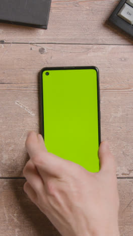 Vertikale-Video-Overhead-Aufnahme-Von-Tisch-Und-Computertastatur-Mit-Der-Hand,-Die-Ein-Green-Screen-Handy-Aufhebt